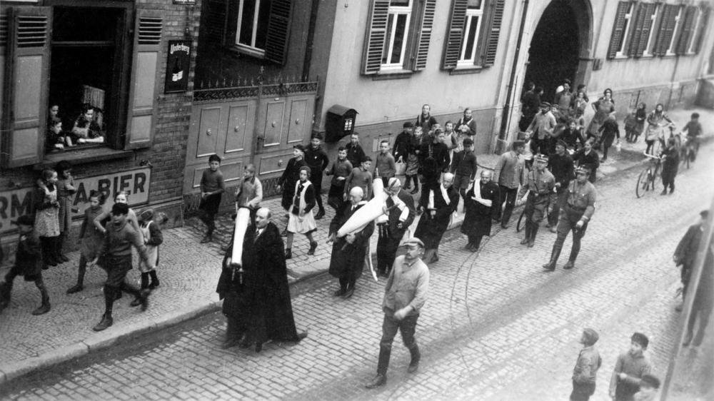 Six Jews are made to walk the streets for hours, wearing prayer shawls and carrying Torah scrolls from the synagogue. They are spit on, scolded, and beaten. The synagogue is looted. Finally, all their belongings are set on fire in the square in front of the town hall.
Guntersblum, 10 November 1938.